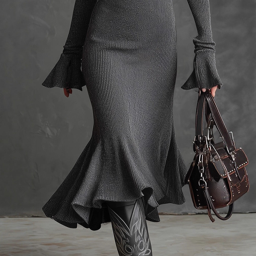 Person wearing a long dark gray knit dress holding a brown leather handbag against a gray background